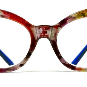 Può includere: Occhiali a gatto con un motivo floreale astratto multicolore nei toni del rosso, rosa, giallo e verde. La montatura ha una finitura lucida con accenti dorati a forma di T sulle cerniere e aste interne di colore blu brillante.