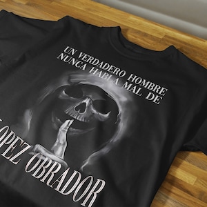 May include: Black t-shirt featuring a white graphic of a skull with a finger over its mouth. The text "UN VERDADERO HOMBRE NUNCA HABLA MAL DE LOPEZ OBRADOR" is printed on the shirt.