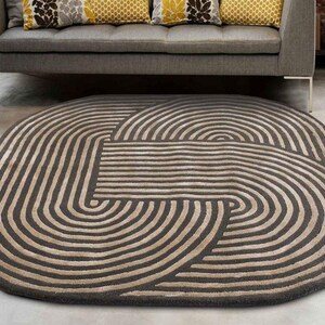 Könnte beinhalten: Ovaler Teppich mit geometrischem Muster. Das Design zeigt geschwungene Linien in Grau- und Beigetönen. Der Teppich liegt auf einem Betonboden, im Hintergrund sind ein graues Sofa und dekorative Kissen zu sehen.