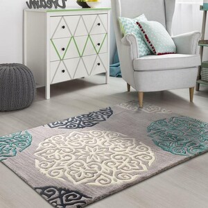 May include: A rectangular gray area rug with large, circular, patterned designs in white, teal, and navy blue. The rug is placed on a light-colored wooden floor, with a gray ottoman and white dresser in the background.