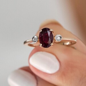 Può includere: Un anello in oro rosa con una grande pietra di rubino ovale e due piccoli diamanti rotondi su entrambi i lati.