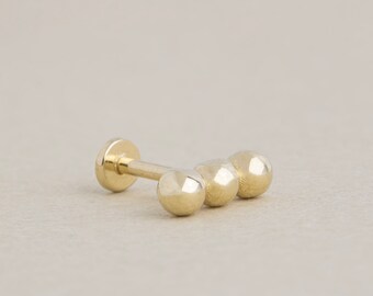 May include: Gold triple ball stud earring. The earring has three round balls in a row, with a post and a flat back.