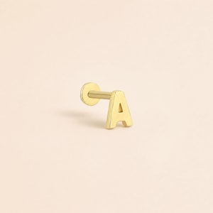 May include: A gold-toned flat back stud earring featuring the capital letter A as the front design. The piece consists of a thin post attached to a circular base, with the letter A positioned at the opposite end of the post.