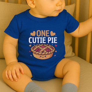 Op de afbeelding: Een blauwe babybody met de tekst "ONE CUTIE PIE" en een cartoon taart. De taart heeft een rasterkorst, roze vulling en een lachend gezicht. De body heeft korte mouwen en een ronde hals.