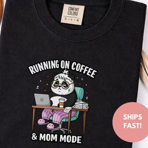 Op de afbeelding: Zwart T-shirt met ronde hals en een afbeelding van een panda met een bril, zittend aan een bureau met een laptop en koffie. De tekst luidt "RUNNING ON COFFEE & MOM MODE". Het shirt heeft een Comfort Colors label.