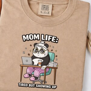 Op de afbeelding: Een beige Comfort Colors t-shirt met een panda met een bril, die op een laptop werkt en een koffiekopje vasthoudt. De tekst luidt "MOM LIFE: TIRED BUT SHOWING UP". De panda zit aan een bureau met boeken.