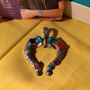 May include: A silver pendant in the shape of a horseshoe with colorful stones. The stones are blue, turquoise, red, yellow, and white. The pendant is on a yellow surface.