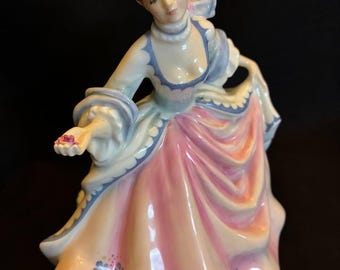 以下が含まれることがあります： 長い流れのあるガウンを着た女性の磁器製の置物。ドレスは主にピンク色で、青と白のアクセントがあります。女性は茶色の髪で、ピンクのリボンをしています。置物は装飾品です。