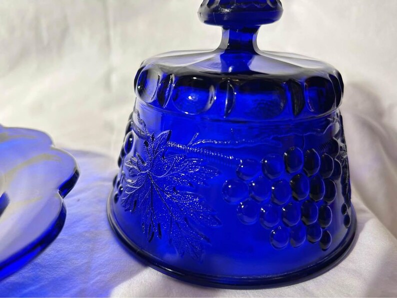 K&ouml;nnte beinhalten: Eine Butterschale aus kobaltblauem Glas mit Deckel. Die Schale ist mit einem Traubenranken- und Traubenmuster verziert.