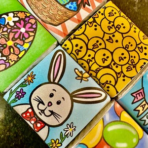 May include: Close-up of several colourful, square-shaped Easter-themed illustrations. Designs include a bunny, chicks, Easter eggs in a basket, and jelly beans. The illustrations are on a dark surface.