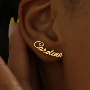 Peut inclure: Boucles d'oreilles dorées avec le nom "Caroline" écrit en cursive. Elles sont portées sur une oreille, touchée par une main. Les ongles sont vernis avec une pointe jaune. Bijou élégant.