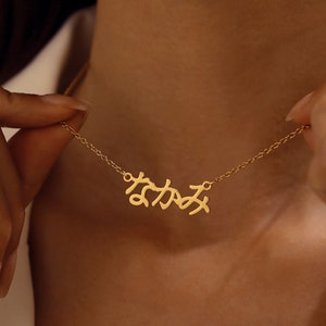 Puede incluir: Un collar de cadena de oro con un colgante de nombre japonés en escritura cursiva. El colgante está hecho de oro y tiene una escritura delicada y fluida.