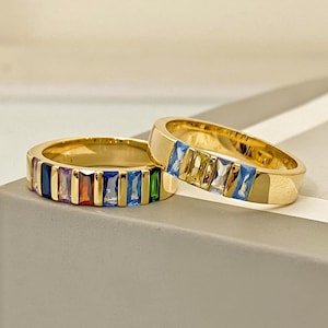 May include: Two gold-colored rings with a row of colorful, rectangular gemstones. The gemstones are in shades of blue, green, yellow, orange, and purple. The rings are on a light gray surface.