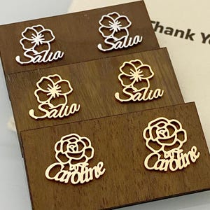 May include: Close-up of several gold-coloured name tags with floral designs. The names "Salia" and "Caroline" are displayed in elegant cursive fonts. The tags are arranged on dark wooden blocks, with a "Thank You" bag in the background.