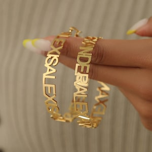May include: Gold-colored hoop earrings with custom names. One earring reads "LEXISALEXIS" and the other reads "KANDERALEIN". The earrings are held up by a person's hand.