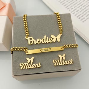 以下が含まれることがあります： ゴールドカラーのジュエリーセット。ネックレス、ブレスレット、イヤリングが含まれています。ネックレスとイヤリングには蝶のアクセントがあります。ネックレスには「Brodie」、ブレスレットには「Chloe」、イヤリングには「Milani」と書かれています。グレーの表面に展示されています。
