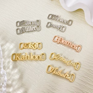 May include: A collection of gold and silver metal shoe charms with various names, including "Kahlo", "Three", "Smooth", "Donte", "Earn", "Miller", "CKJ", "La Moto", and "Nathalie".