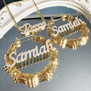 Puede incluir: Dos pendientes de aro dorados con el nombre "Samiah" escrito en brillantes de imitación. Los pendientes son de estilo bambú. También se muestra un collar de cadena dorada con el nombre "Drianna" escrito en brillantes de imitación.