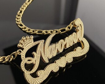 May include: Gold-tone nameplate necklace with the name "Naomi" in a cursive font with a crown above the first letter. The necklace has a curb chain.