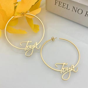 Könnte beinhalten: Ein Paar goldfarbene Creolen. Jeder Ohrring trägt den Namen "Toya" in Schreibschrift. Die Ohrringe werden auf einer hellen Oberfläche präsentiert, mit gelben Blumen und dem Text "FEEL NO RE" auf einer Schachtel im Hintergrund.