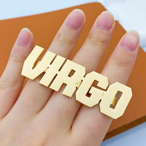 May include: A gold-colored ring spelling out the word "VIRGO" in bold, block letters. The ring is shown on a finger, with a brown surface in the background. The ring is a statement piece, likely for astrology enthusiasts.