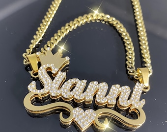 May include: Gold chain necklace with a pendant featuring the name "Sianni" spelled out in cursive with rhinestones. The pendant also has a small crown and a heart with rhinestones.