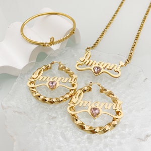 May include: Gold necklace, bracelet, and hoop earrings with the name "Immani" and a pink heart-shaped stone.