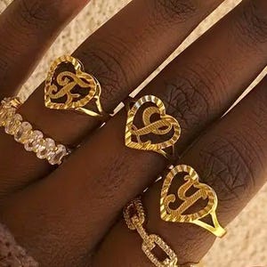 Peut inclure: Une main ornée de plusieurs bagues en or. Les bagues comprennent des motifs en forme de cœur avec des lettres, une bague de style chaîne et une bague avec des pierres transparentes. Les ongles sont longs et peints avec un dégradé d'orange et de blanc.