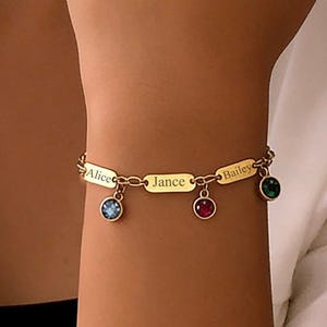 Puede incluir: Pulsera dorada con tres placas con nombres y dijes de piedras preciosas. Las placas dicen "Alice", "Jance" y "Bailey". Los dijes son azules, rojos y verdes. La pulsera se lleva en la muñeca.