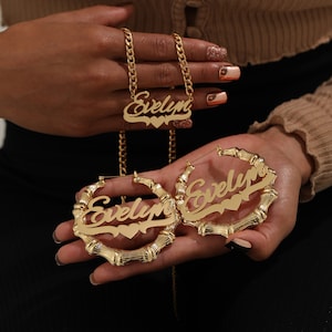 May include: Gold-colored jewelry set featuring a necklace and hoop earrings. The necklace has a chain with the name "Evelyn" in a cursive font, with a heart detail. The hoop earrings also display the name "Evelyn" in a similar style.