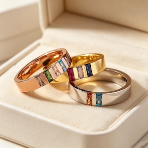 May include: Three metal rings featuring baguette-cut gemstones in various colors. One ring is rose gold with light blue, green, and clear stones. Another is yellow gold with pink, white, and blue stones. The third is silver with orange and blue stones.