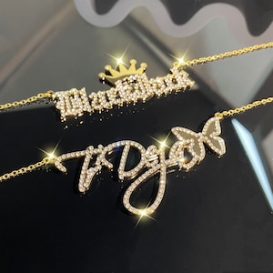 May include: Two gold necklaces with a crown and butterfly charm. The necklaces are adorned with sparkling rhinestones and spell out the names "Maverick" and "T'Dajah".