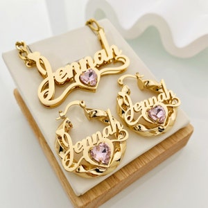 May include: Gold-tone necklace and earrings set with the name "Jenna" spelled out in cursive with a pink heart-shaped rhinestone in the center of each piece.