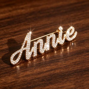 May include: A gold-toned brooch spelling "Annie" in a cursive font, encrusted with sparkling crystals. The pin is set against a dark wood surface, creating a contrast that highlights the brooch's design. The brooch is a personalized accessory.