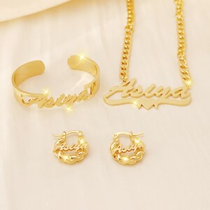 May include: Gold-toned jewelry set with the name "Asliya" engraved on a necklace, bracelet, and earrings. The necklace has a curb chain and a heart charm. The bracelet is a simple band. The earrings are small hoops.