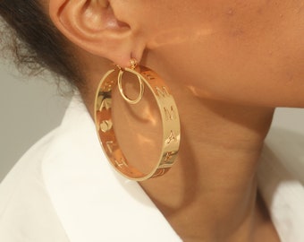 May include: Gold hoop earrings with a cutout design featuring the letters "EMMA" around the hoop. The earrings are worn by a person with dark skin.