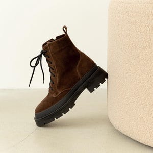 Puede incluir: Una bota de combate de ante marrón con cordones negros y una suela negra gruesa. La bota tiene un tobillo alto y una lengüeta en la parte trasera. La bota está apoyada contra un objeto beige, texturizado y redondeado.