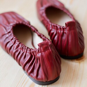 May include: A pair of red leather ballet flats with a gathered detail on the top. The flats are on a light wood surface.