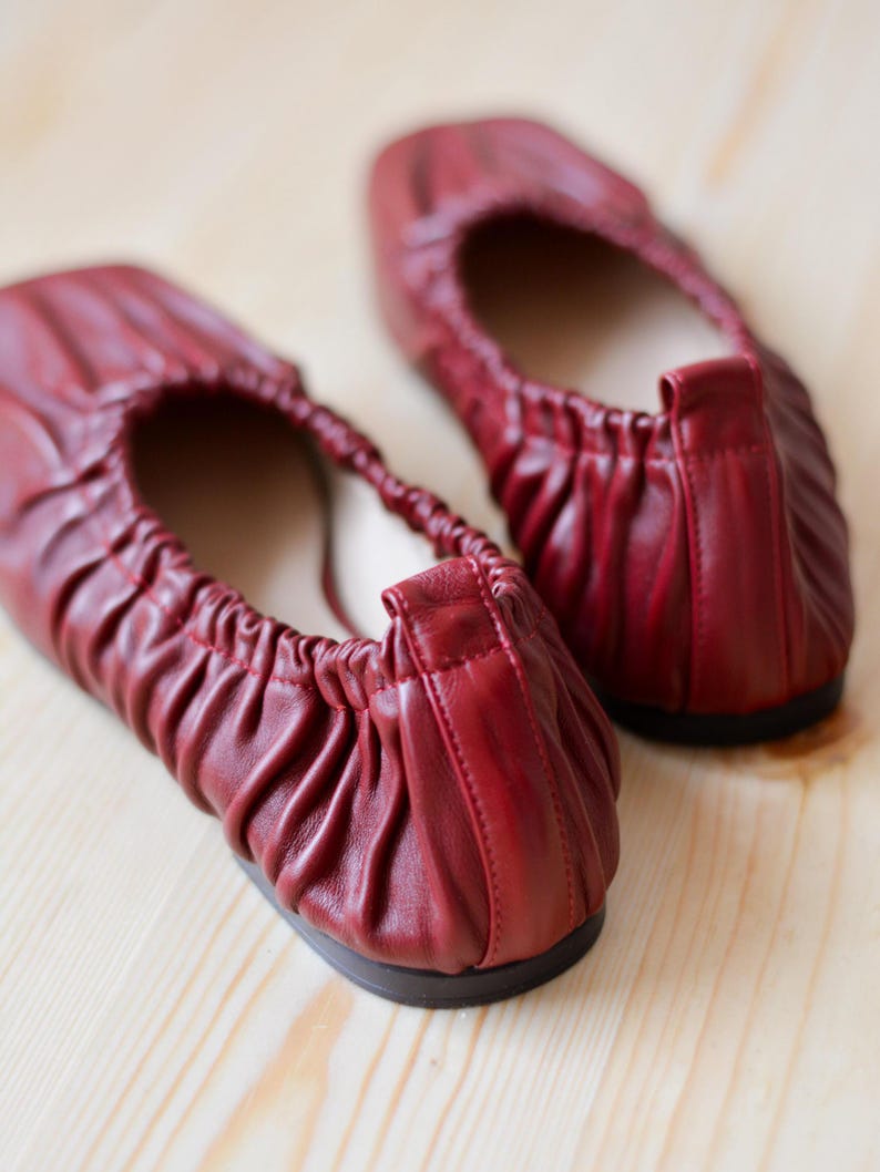 May include: A pair of red leather ballet flats with a gathered detail on the top. The flats are on a light wood surface.