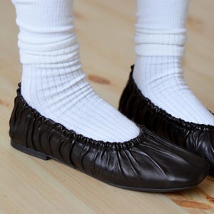 May include: A pair of black leather ballet flats with a gathered detail around the ankle. The flats are worn with white socks.