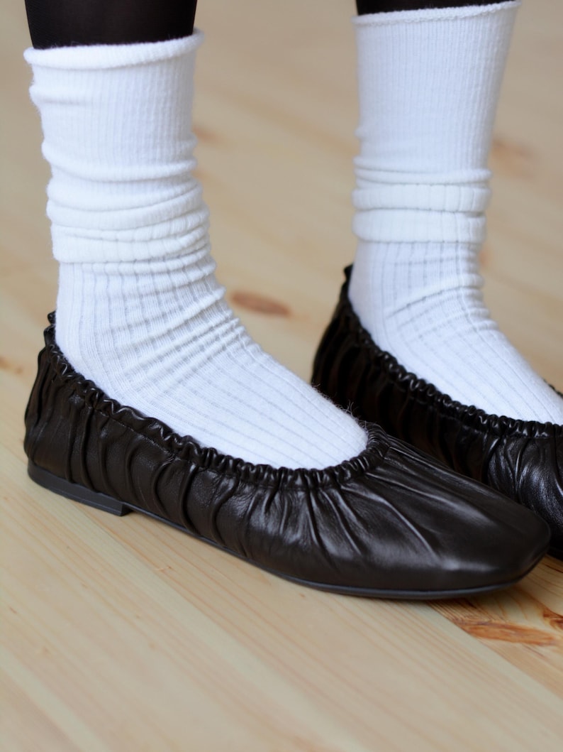 May include: A pair of black leather ballet flats with a gathered detail around the ankle. The flats are worn with white socks.