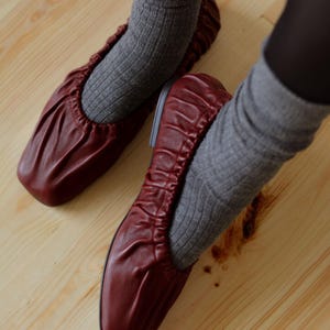 May include: A pair of burgundy leather ballet flats with a gathered, scrunched design. The flats are worn with gray socks.