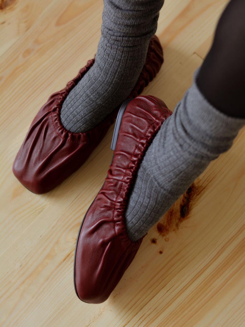 May include: A pair of burgundy leather ballet flats with a gathered, scrunched design. The flats are worn with gray socks.