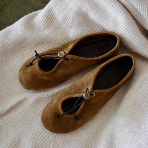 May include: A pair of brown suede ballet flats with a round toe and a small opening on the top. The flats have a black drawstring closure with a silver-colored metal accent. The shoes are resting on a white, textured fabric.