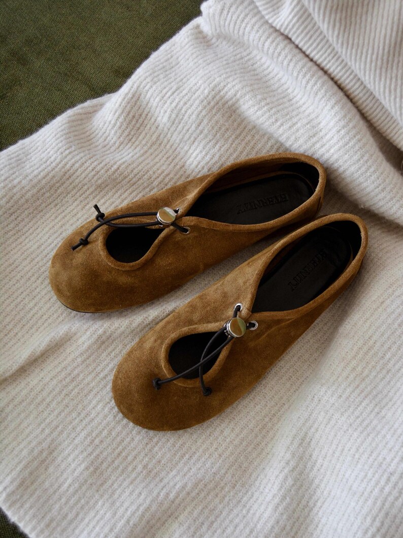 May include: A pair of brown suede ballet flats with a round toe and a small opening on the top. The flats have a black drawstring closure with a silver-colored metal accent. The shoes are resting on a white, textured fabric.