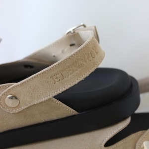May include: Beige suede sandals with a black rubber sole and a buckle closure. The strap has the text "BIRKENSTOCK" embossed on it.