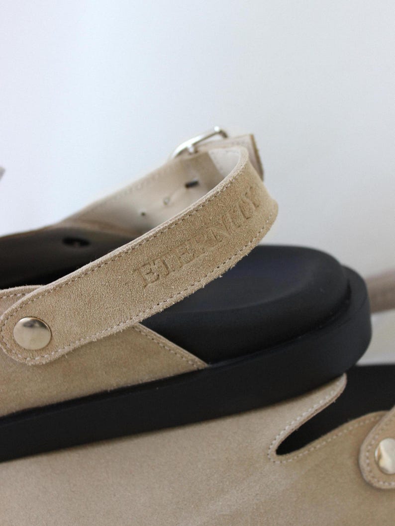 May include: Beige suede sandals with a black rubber sole and a buckle closure. The strap has the text "BIRKENSTOCK" embossed on it.