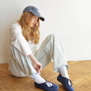 May include: A person wearing a blue cap, white long-sleeve shirt, and light blue jeans, sitting on a wooden floor. The person is wearing navy blue flats with a tie detail. The flats are a classic style, suitable for casual wear.