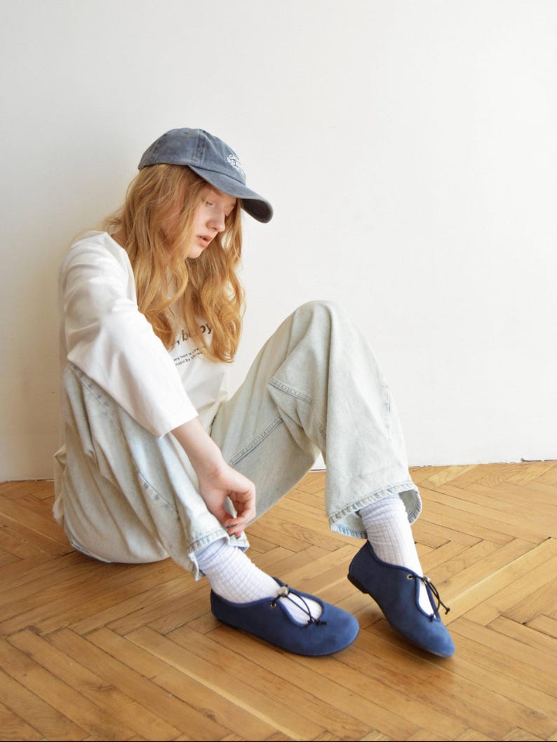 May include: A person wearing a blue cap, white long-sleeve shirt, and light blue jeans, sitting on a wooden floor. The person is wearing navy blue flats with a tie detail. The flats are a classic style, suitable for casual wear.