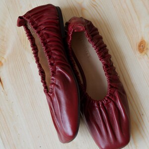 May include: A pair of burgundy leather ballet flats with a gathered, scrunched design. The flats are on a light wood surface.